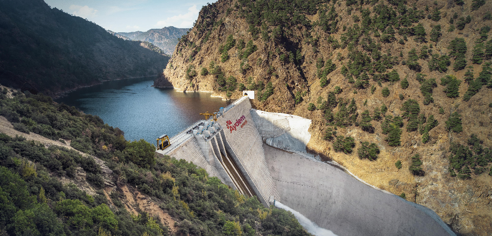 Türkiye’nin öncü enerji şirketi olan Aydem’in portföyünde yer alan hidroelektrik santralinin bulunduğu bölgedeki fotoğrafıdır. Fotoğraf, geniş açıdan çekilmiş, ağaçlık, dağlık ve su kaynağının başında duran hidroelektrik santrali içermektedir.